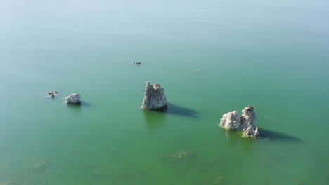 Aerial-shot-of-Mono-Lake's-tufa-towers-set-in-calm-blue-waters,-highlighting-the-unique-geological-formations-and-expansive-surrounding-landscape-under-a-sunny-sky