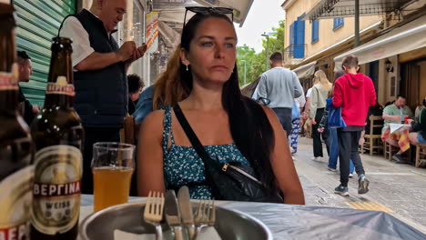 Young-woman-sitting-at-table-on-busy-street,-foot-traffic,-people-moving-in-Athens-Greece