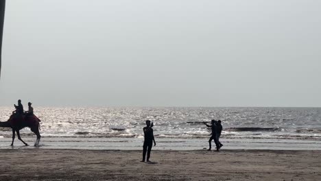 Touristen-Reiten-Und-Reiten-Kamele-Am-Strand-Mit-Der-Meeresbrise-Hinter-Ihnen