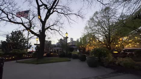 An-evening-scene-in-a-park-with-trees-adorned-with-string-lights,-an-American-flag-fluttering,-and-the-backdrop-of-city-buildings-under-the-Brooklyn-Bridge