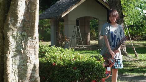 Youthful,-relaxed-Asian-teenage-girl-stops-to-admire-lush-garden-bushes-and-flowers-while-carrying-acoustic-guitar-during-daytime