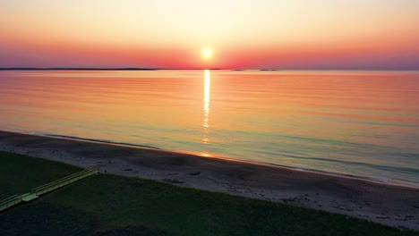 Aerial-Drone-Rising-Over-Wooden-Walking-Path-Leading-to-Colorful-Ocean-Sunrise-on-Beach-with-Trees-and-Grass-at-Dawn-with-Beautiful-Red,-Orange,-and-Purple-Colors-in-the-Sky-and-Reflecting-off-Water