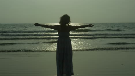 A-silhouette-woman-stands-on-a-beach,-arms-outstretched,-with-the-sun-setting-in-the-background