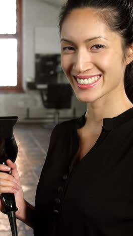 Smiling-female-hairstylist-holding-hair-dryer-in-hair-salon