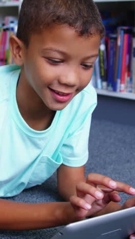 Schoolboy-lying-on-floor-and-using-digital-tablet-in-library-at-school