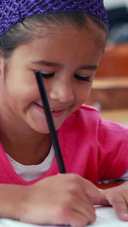 Cute-little-girl-colouring-in-book-in-classroom-smiling-at-camera