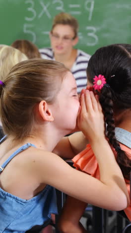 Schoolgirl-whispering-into-her-friend-s-ear-in-classroom