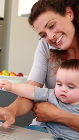 Mutter-Sitzt-Mit-Baby-Auf-Dem-Schoß,-Benutzt-Laptop-Und-Telefoniert