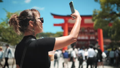 Una-Mujer-Vestida-De-Negro-Ajusta-Sus-Gafas-De-Sol-Mientras-Está-De-Pie-Bajo-Una-Puerta-Torii-Roja-En-Un-Santuario-Japonés