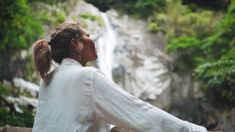 Una-Mujer-Asiática-Se-Encuentra-Al-Borde-De-Una-Cascada,-Admirando-El-Espectáculo-Natural,-Rodeada-De-Exuberante-Vegetación.