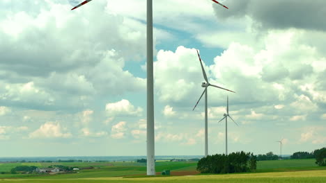 Aerial-footage-highlighting-a-row-of-wind-turbines-standing-tall-over-green-agricultural-fields-under-a-bright,-partly-cloudy-sky