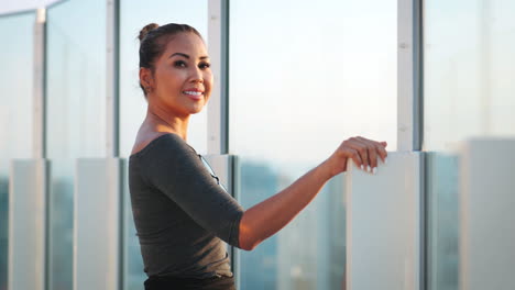 Asian-female-model-poses-at-a-glass-railing,-enjoying-the-urban-skyline-view