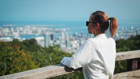 Woman-with-sunglasses-and-white-shirt-gazes-over-a-coastal-city-from-a-wooden-railing