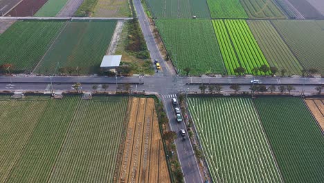 Aerial-drone-flyover-soaring-above-agricultural-farmlands,-captures-a-picturesque-rural-landscape,-with-traffic-converging-at-the-quaint-country-crossroads,-showcasing-the-beauty-of-the-countryside