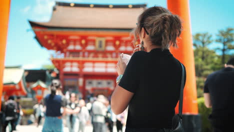 Woman-enjoying-iced-coffee-beside-an-orange-torii-gate,-wearing-sunglasses-and-tied-up-hair