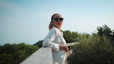 Stylish-woman-in-white-shirt-leans-against-a-wooden-railing,-with-a-city-and-ocean-backdrop,-greenery-around-and-confident-pose-emphasize-exploration-and-enjoying-nature's-beauty-alongside-urban-life