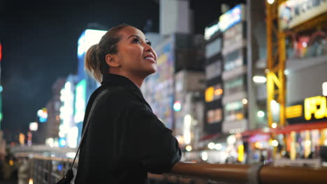 Una-Mujer-Vestida-De-Negro-Se-Encuentra-En-Un-Puente,-Contemplando-Un-Vibrante-Paisaje-Urbano-Lleno-De-Luces-De-Neón-Y-Edificios-Altos