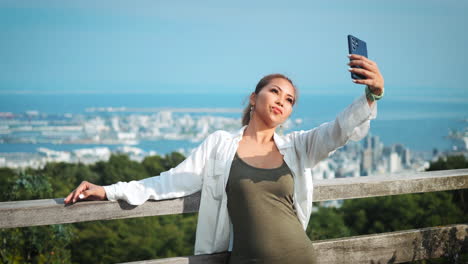 Young-woman-with-dark-blonde-hair-in-a-ponytail-takes-a-selfie-with-a-smartphone-against-a-backdrop-of-a-sprawling-city-and-blue-sky