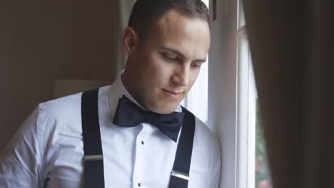 Groom-looks-up-happily-while-leaning-against-window