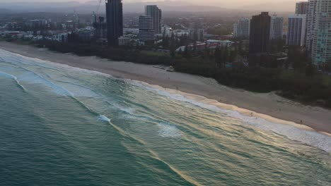 Eine-Luftaufnahme-Aus-Der-Vogelperspektive-Zeigt-Den-Langen,-Ausgedehnten-Sandstrand-Mit-Wellen,-Die-Gegen-Das-Ufer-Schlagen,-Luxuriöse-Apartments-Am-Wasser-Und-Resorthotels-Mit-Meerblick-An-Der-Goldküste-Von-Broadbeach-Bei-Sonnenuntergang