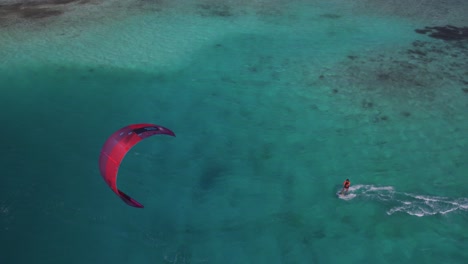 Una-Persona-Practicando-Kitesurf-Con-Una-Cometa-Roja-Sobre-Aguas-Cristalinas-De-Color-Turquesa-En-Los-Roques,-Vista-Aérea