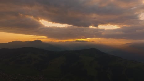 Aerial-view-showing-an-orange-sunset-as-it-casts-a-warm-glow-between-a-cloudy-sky-and-distant-mountain-ranges,-sun-rays