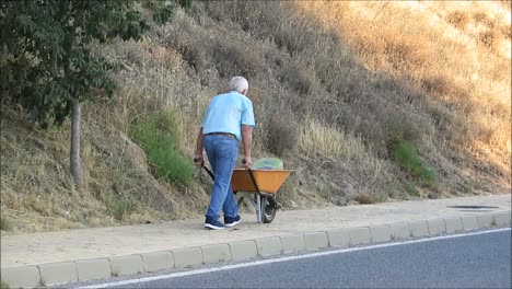 Álora,-España---25-De-Agosto-De-2018:-Anciano-Empujando-Una-Carretilla-Con-Un-Saco-De-Cemento-Por-Una-Acera-Rural