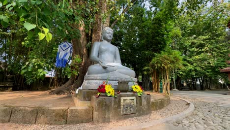 Nahaufnahme-Einer-Buddha-Statue-Mit-Blumen-Auf-Dem-Sockel-Unter-Einem-Baum