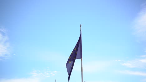 Europe-Union-flag-waving-on-high-pole-with-palm-trees-bellow,-tilt-up-view