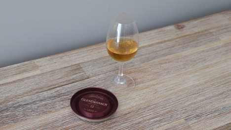 Tapa-Metálica-Giratoria-Del-Embalaje-De-Una-Botella-De-Whisky-Escocés-De-Pura-Malta-Original-Glendronach-De-Doce-Años,-Junto-A-Un-Vaso-De-Copita-Lleno-De-Whisky,-Sobre-Una-Superficie-De-Madera