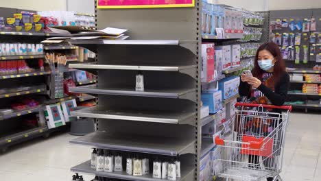 A-shopper-wears-face-mask-stands-next-to-empty-supermarket-shelves,-usually-stocked-with-toilet-paper