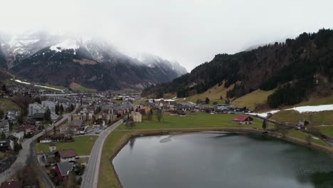 Aerial-View-of-Engelberg-Town-and-Alpine-Lake-Under-Snow-Capped-Peaks-of-Swiss-Alps,-Establishing-Drone-Shot