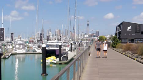 Couple-walking-on-a-wooden-path-dock-at-Westhaven,-Auckland-skyline-in-background,-New-Zealand