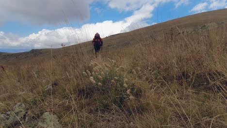 Low-angle:-Young-male-backpacker-walks-toward-camera-in-hilly-meadow