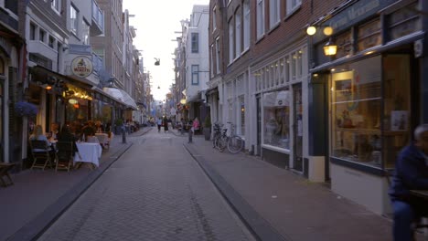 Forward-moving-dolly-shot-of-a-street-in-Amsterdam,-The-Netherlands,-where-people-are-enjoying-themselves-on-terrasses-and-in-restaurants-in-the-early-evening