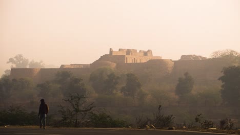 A-south-asian-villager-walking-by-the-road-with-an-Ancient-Indian-fort-or-castle-in-background-in-a-village-during-morning-time