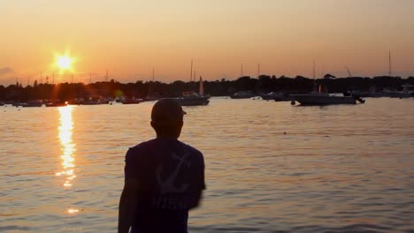 Hombre-Saltando-Una-Roca-Hacia-El-Puerto-Lleno-De-Barcos-Al-Atardecer