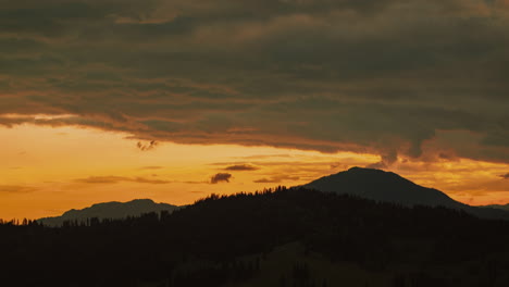 Zeitraffer-Zeigt-Einen-Orangefarbenen-Sonnenuntergangshimmel,-Der-Einen-Warmen-Schein-Zwischen-Einem-Bewölkten-Himmel-Und-Entfernten-Berggipfeln-Wirft