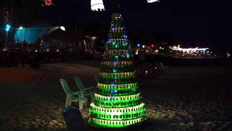 A-beer-bottle-shaped-Christmas-tree-on-a-Thai-beach-flickering-its-lights-at-night,-shot-panning-left