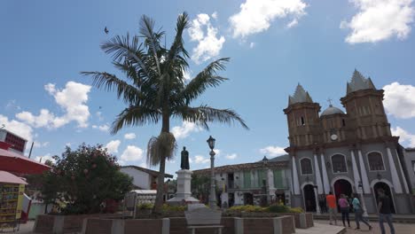 Replica-of-old-colonial-church-in-sunny-plaza-with-palm-tree-and-statues,-Colombia