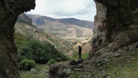 Woman-stands-under-massive-rock-arch-looking-over-hilly-landscape