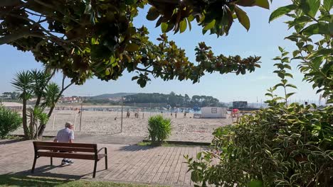 Un-Anciano-Hace-Ejercicios-De-Estiramiento-En-Un-Banco-En-La-Acera-Frente-A-La-Playa-Con-Gente-Practicando-Deportes-Y-Volea-En-La-Arena,-Un-Día-Soleado,-Tiro-Inclinado