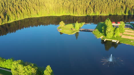 Vista-Aérea-De-Un-Pintoresco-Lago-Con-Fuente-Y-Un-Entorno-Verde-En-Una-Puesta-De-Sol-Dorada