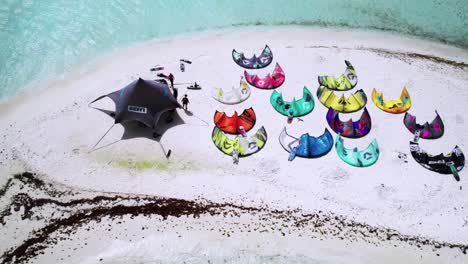 A-vibrant-kite-camp-setup-on-a-tiny-sandy-island-surrounded-by-turquoise-waters,-aerial-view