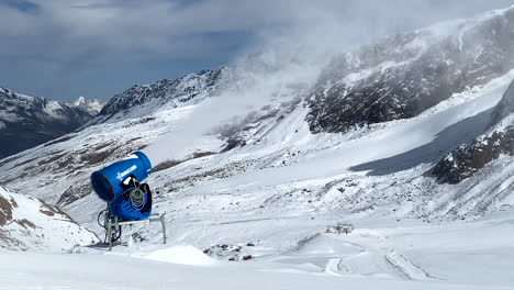 Una-Máquina-De-Nieve-Azul-En-Pleno-Funcionamiento-Que-Sopla-Nieve-Artificial-Blanca-En-Las-Laderas-De-Una-Popular-Estación-De-Esquí,-Las-Laderas-Rodeadas-Por-Hermosas-Montañas-Del-Glaciar-Val-Senales,-Italia