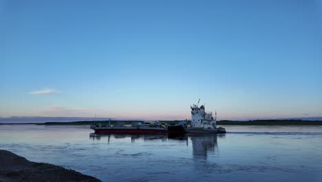 Ein-Binnenschiff-Wird-Bei-Sonnenuntergang-Von-Einem-Schlepper-Einen-Fluss-Hinunter-Geschleppt