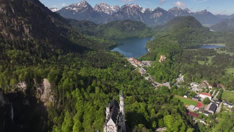 Vista-Aérea-Del-Castillo-De-Neuschwanstein-En-Schwangau,-Alemania,-Con-Una-Cadena-Montañosa-Y-Una-Zona-Residencial-Al-Fondo,-Capturando-El-Concepto-De-Arquitectura-De-Cuento-De-Hadas-Y-Belleza-Escénica-Alpina