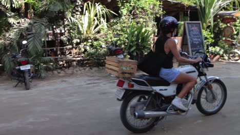 Young-girl-riding-a-motorcycle-on-a-sunny-day-on-a-muddy-dirt-road-in-a-laidback-hippy-town-in-Mexico,-slow-motion