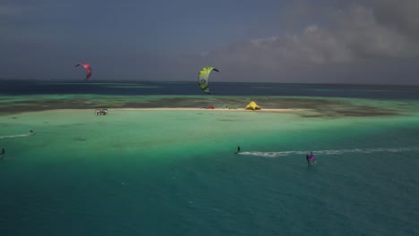 Kiteboarders-navigating-clear-blue-waters-near-a-tropical-island-under-cloudy-skies,-aerial-view