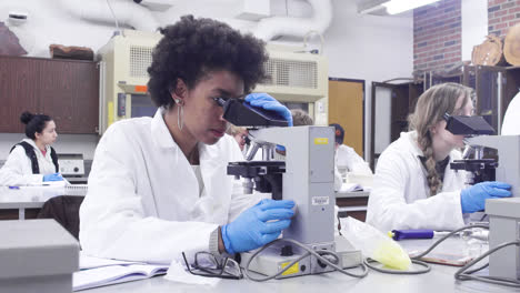 A-black-woman-college-student-biology-major-in-white-lab-coat-and-gloves-researches-with-a-microscope-at-California-State-University-Chico-slow-motion-4k